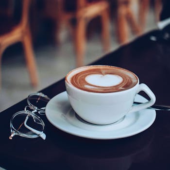 Aesthetic photo of a cappuccino with heart foam, accompanied by eyeglasses on a table.